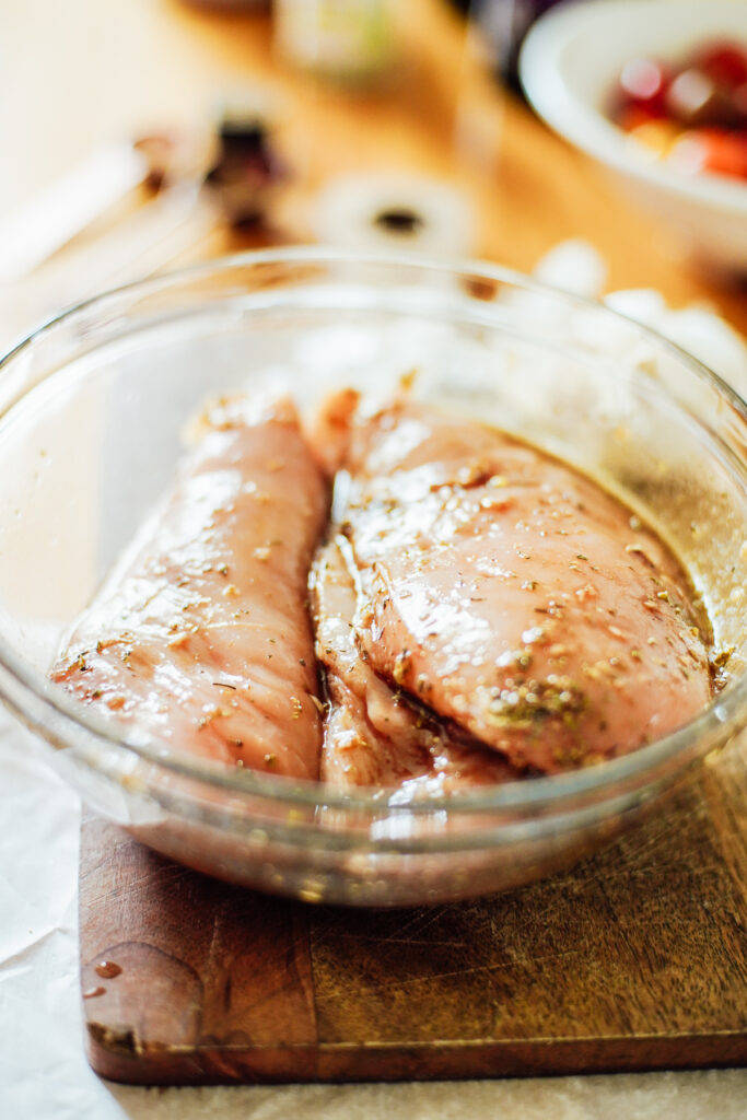 Chicken breasts marinating in a glass bowl with olive oil, balsamic vinegar, Italian seasoning, and garlic.