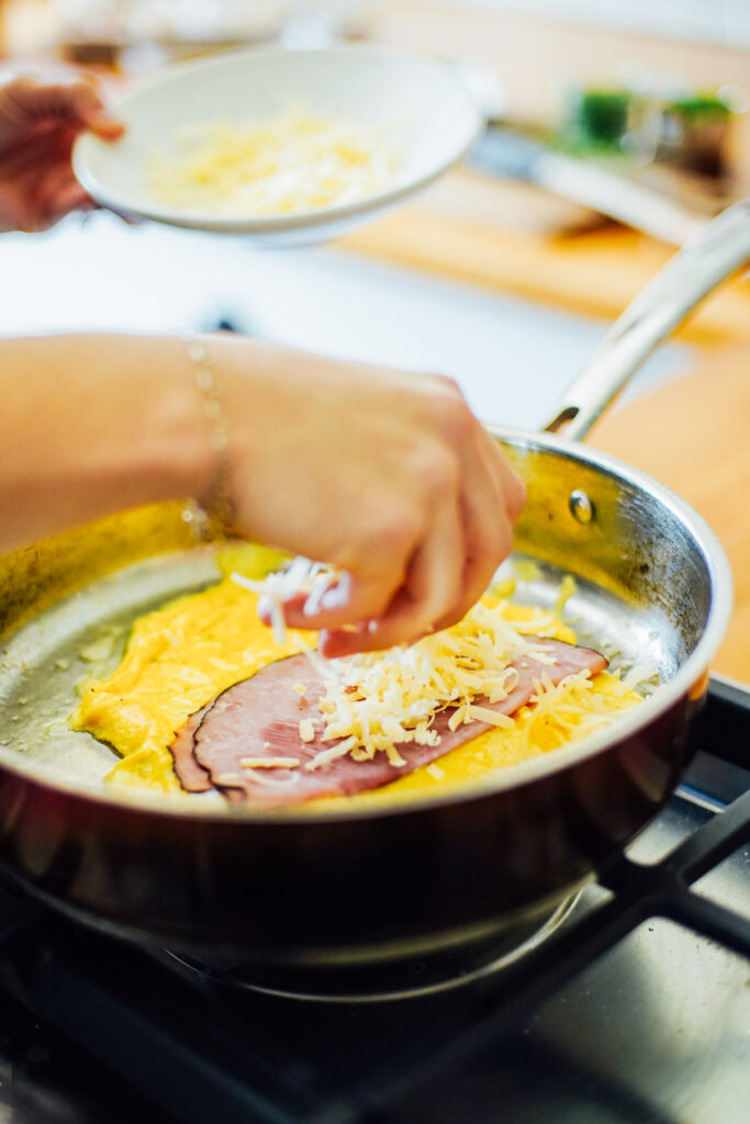Adding shredded cheese and ham to the omelette while cooking in a skillet.