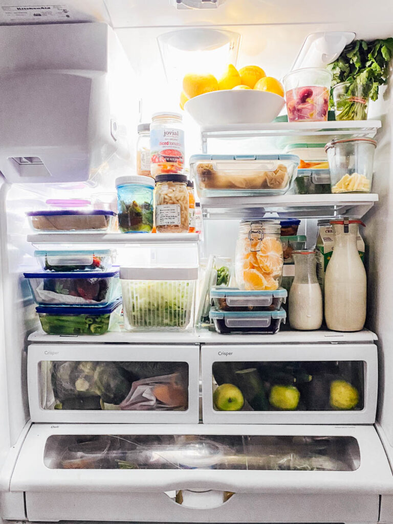 Open fridge full of fresh ingredients, including fresh herbs in glass of water.