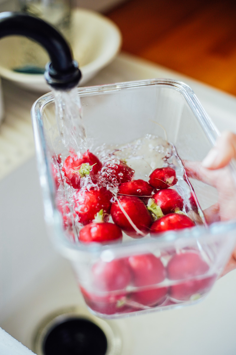 Filling a large glass bowl with radishes inside with fresh water at the sink.