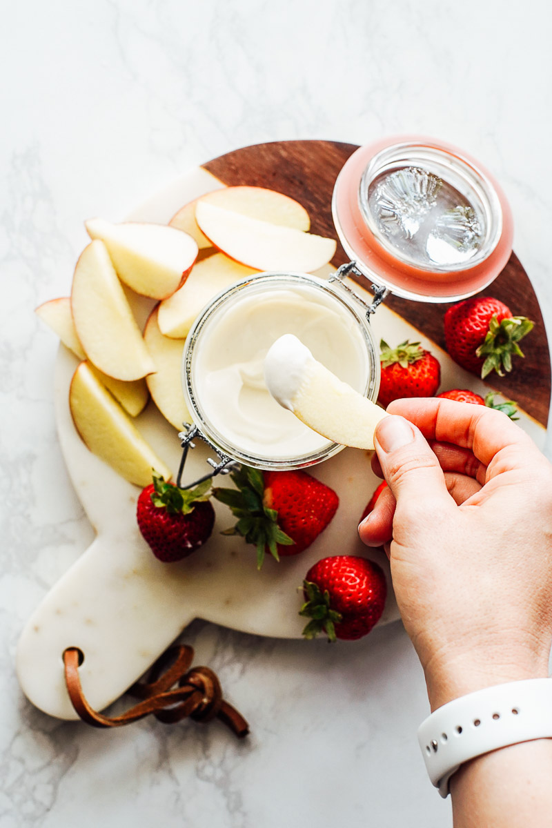 Dipping a sliced apple into yogurt dip on a cutting board with apples and strawberries spread around.