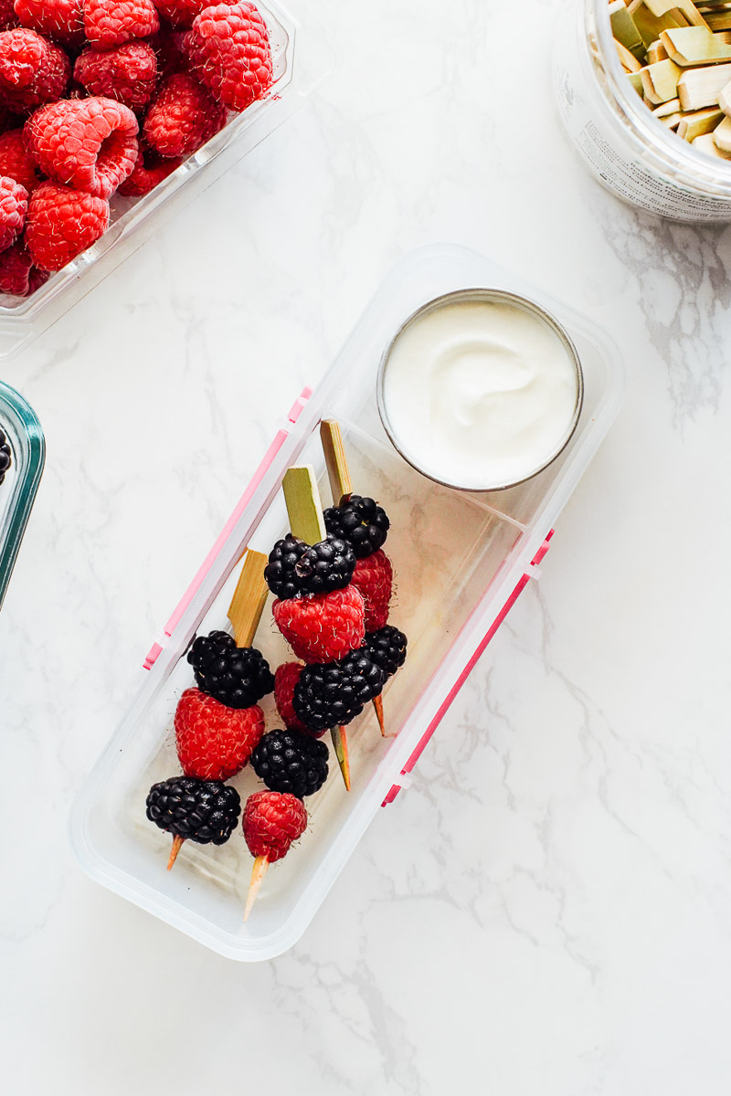 Yogurt fruit dip in a to-go container with raspberries and blackberries on a kabob stick.