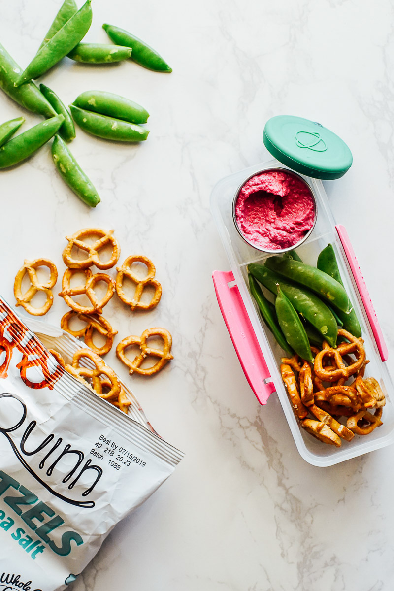 Homemade hummus, pretzels, and snap peas in a plastic snack container.