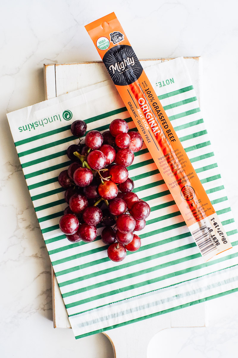 Mightie Sticks brand beef jerky on a cutting board with grapes.