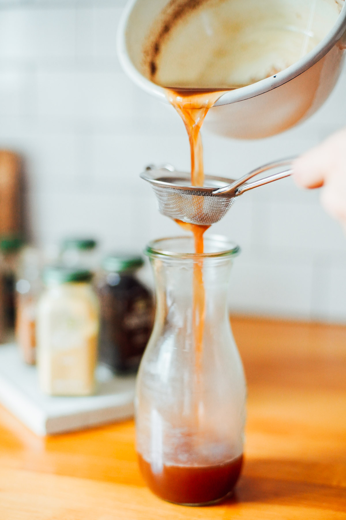 Pouring the chai concentrate through a fine mesh sieve into a tall glass bottle.