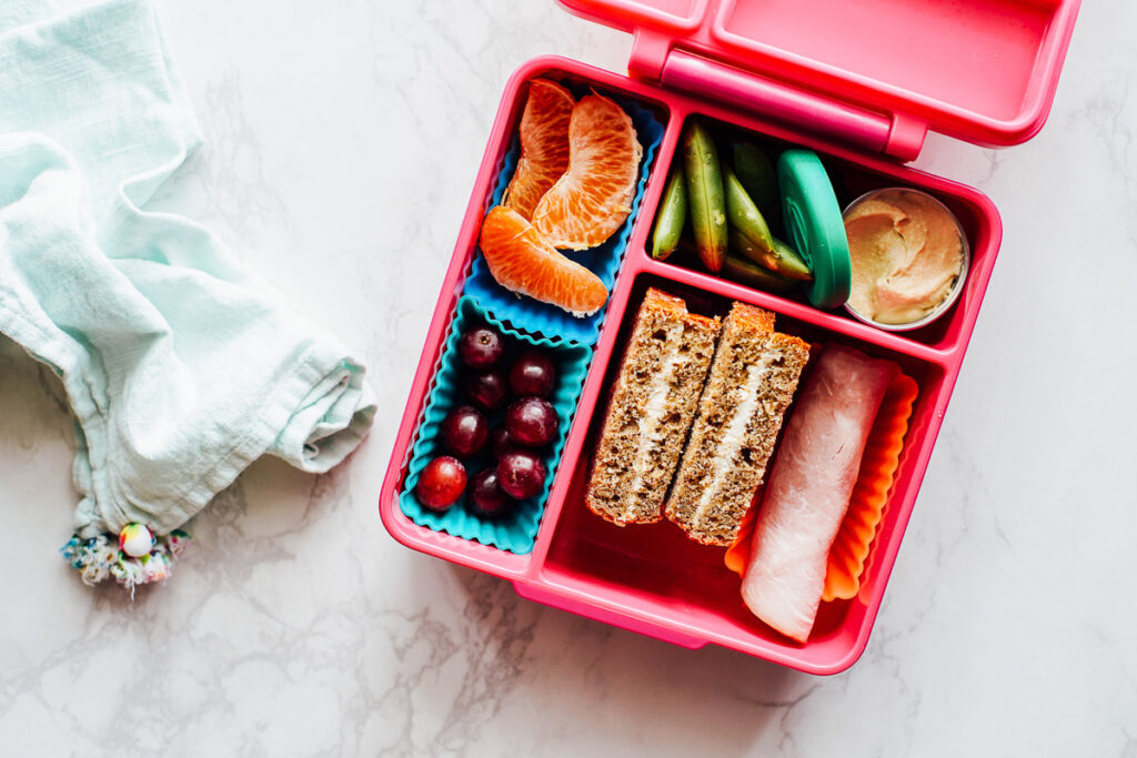 Pink bento box with a banana bread cream cheese sandwich and rolled up ham, grapes, oranges, hummus, and snap peas.