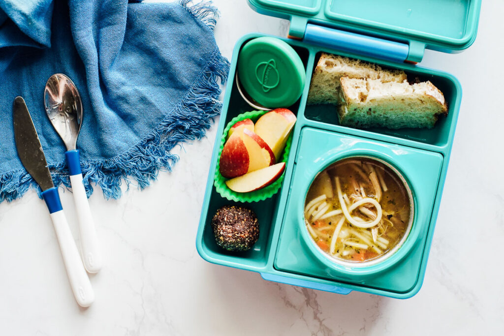 Chicken noodle soup, energy bite, apple slices, and bread in a bento box lunchbox.