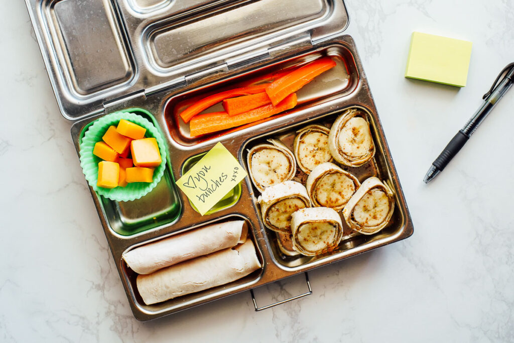 Banana rolled up in a tortilla and peanut butter, carrot sticks, cheese cubes, and turkey rolled up in a lunchbox.