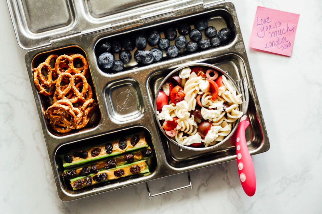 Pasta salad in a lunchbox with celery and peanut butter, pretzels, and blueberries.