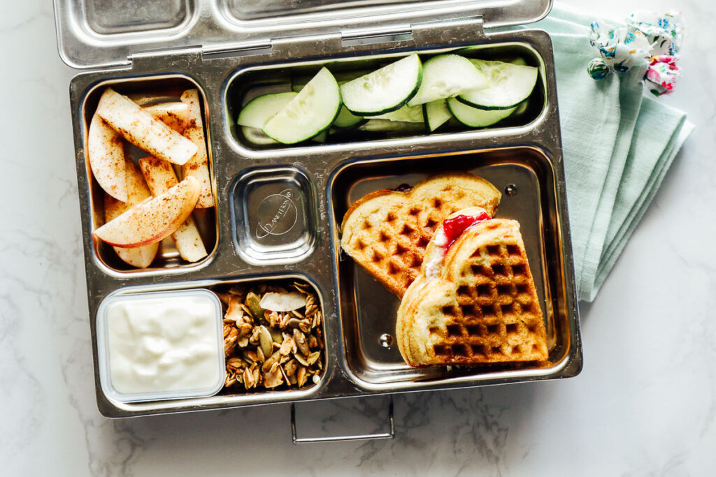 Waffle sandwich (with cream cheese and jam), yogurt and granola, apple slices, and cucumbers in a lunchbox.