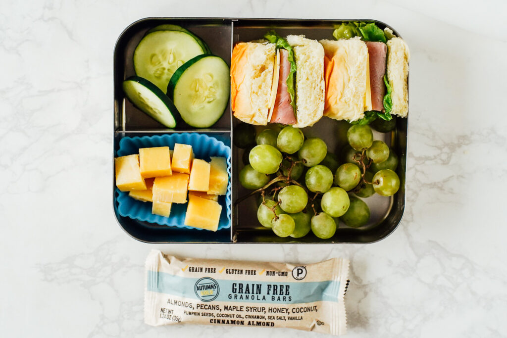 Ham and cheese sandwich on dinner rolls with grapes, cheese cubes, and cucumber slices in a lunchbox.