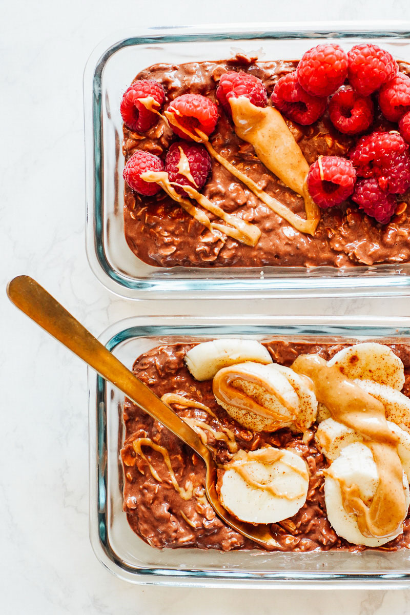 Chocolate overnight oats in glass meal prep bowls, topped with bananas and raspberries.