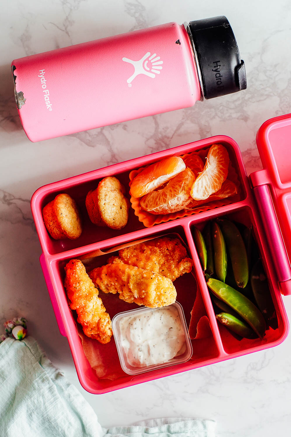 Chicken nuggets, mini muffins, sugar snap peas, orange slices, and ranch dip in a lunchbox.