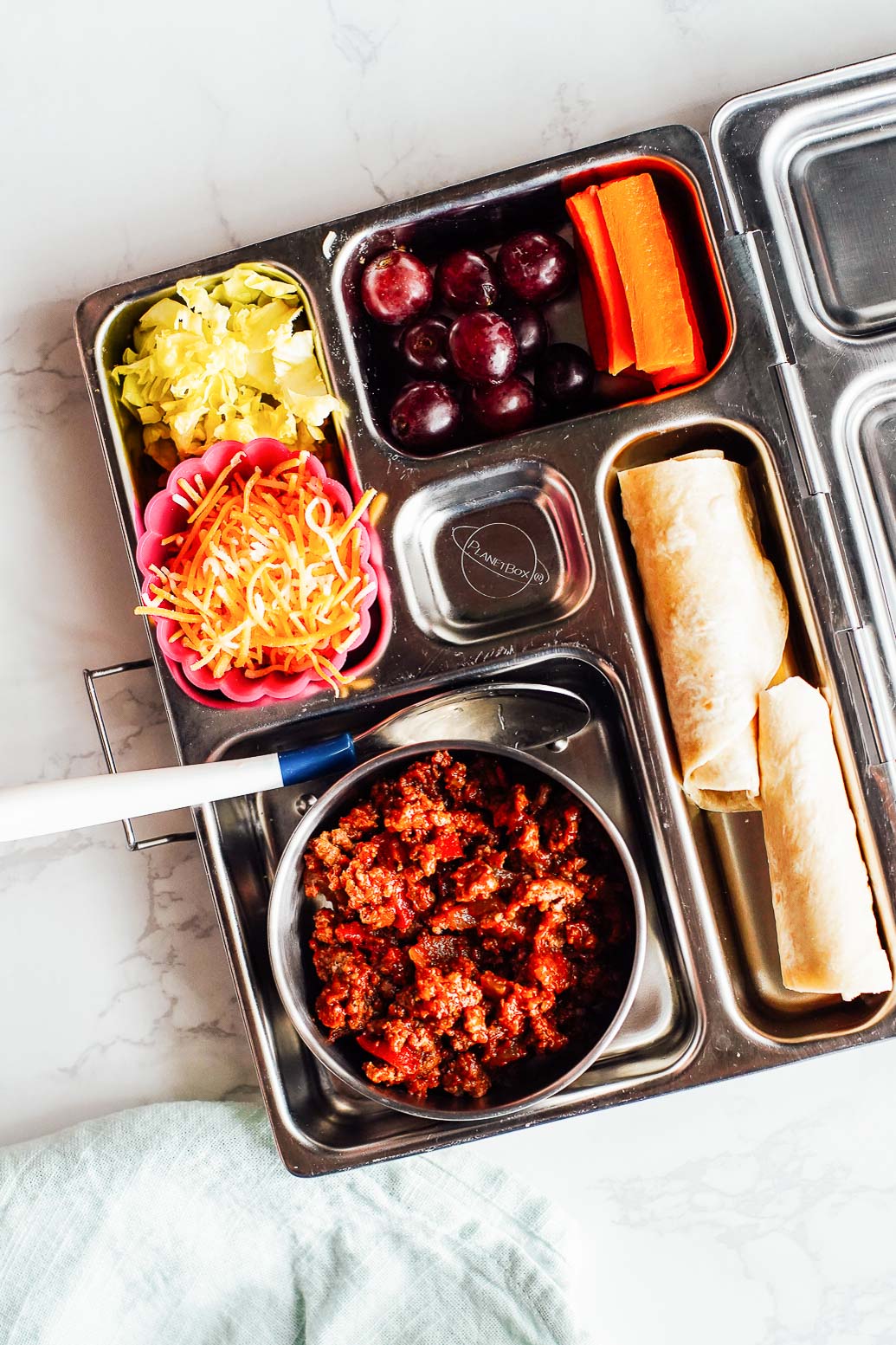 Build your own tacos: leftover ground beef taco meat with a spoon, tortilla on the side, shredded cheese, lettuce, grapes, and carrots in a bento box.