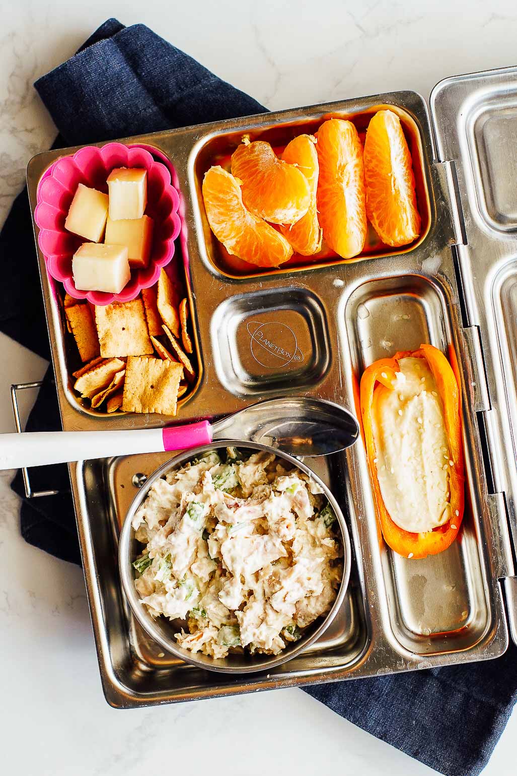 Tuna salad with crackers and cheese, orange slices, and a mini pepper with hummus in a bento box lunchbox.