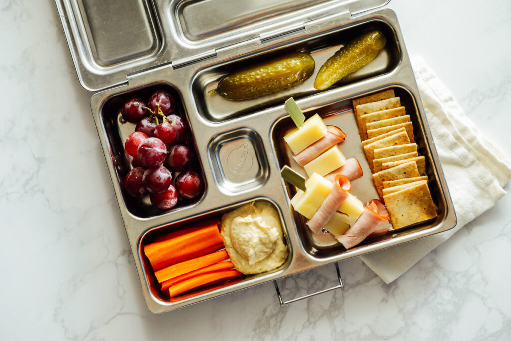 Homemade cheese, crackers, and ham lunchable with carrots, hummus, grapes, and a pickle.