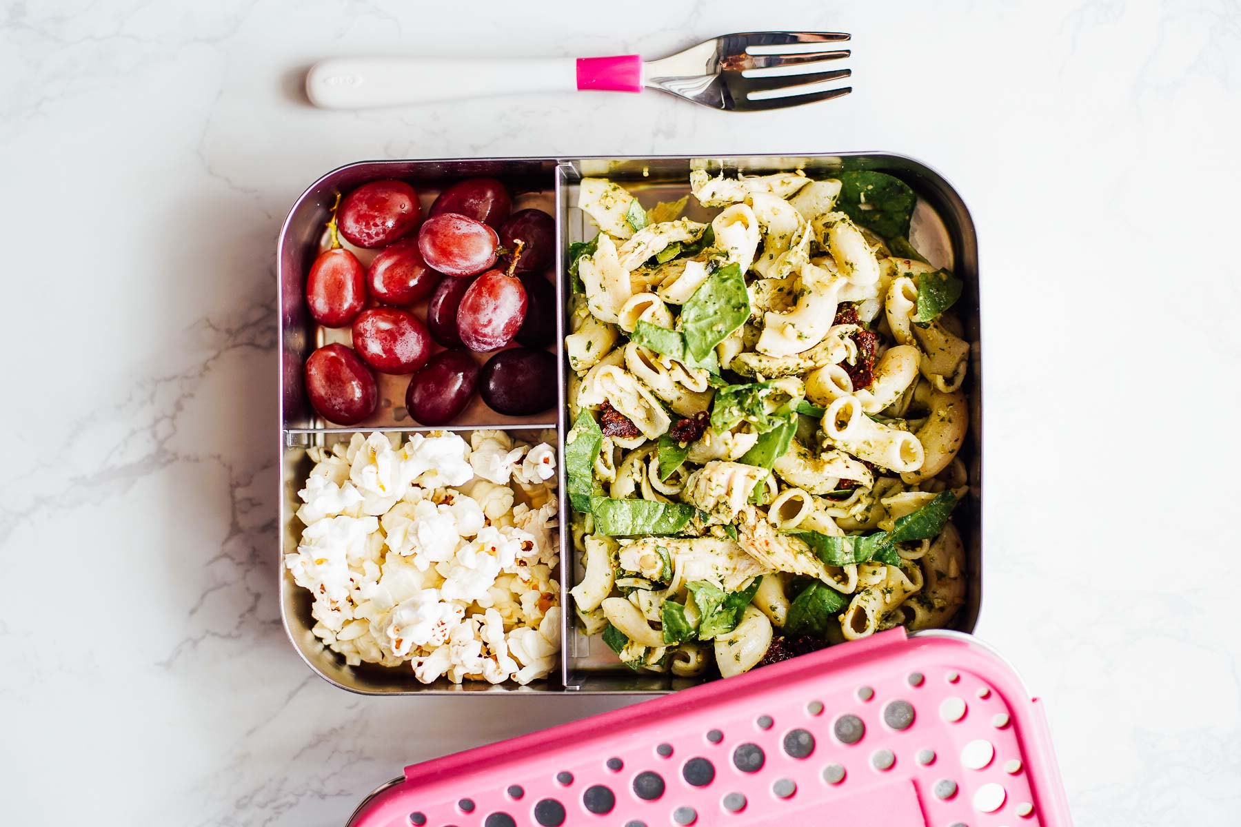 Pasta salad in a lunchbox with popcorn and grapes.