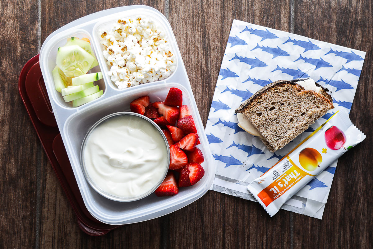 Yogurt and strawberries, popcorn, cucumber slices, turkey sandwich on sourdough bread, and a Just fruit bar in a lunchbox. 