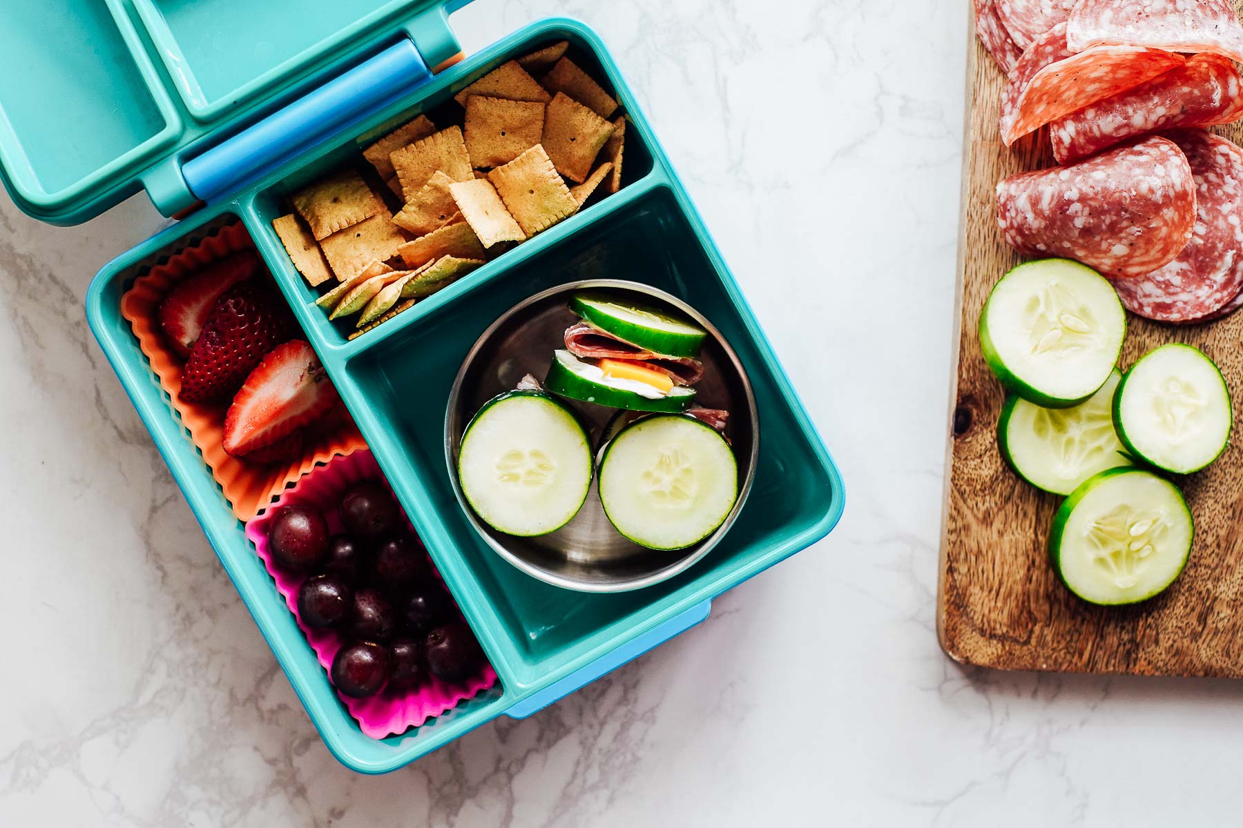 Cucumber slices with cheese and cream cheese and salami, crackers, grapes, and berries in a lunchbox.