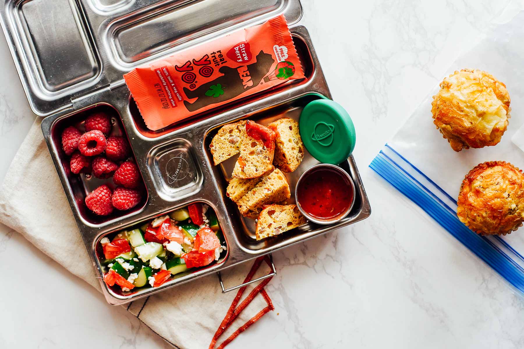 Pizza Muffins with dipping sauce, salad, raspberries, and fruit roll up in a lunchbox.