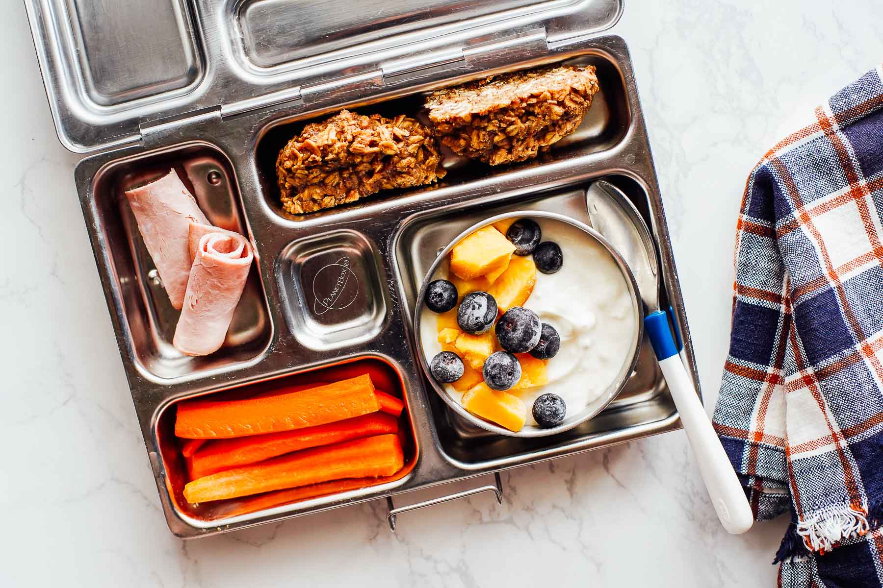Yogurt with fruit, carrots, ham roll ups, and breakfast cookie in a lunchbox.