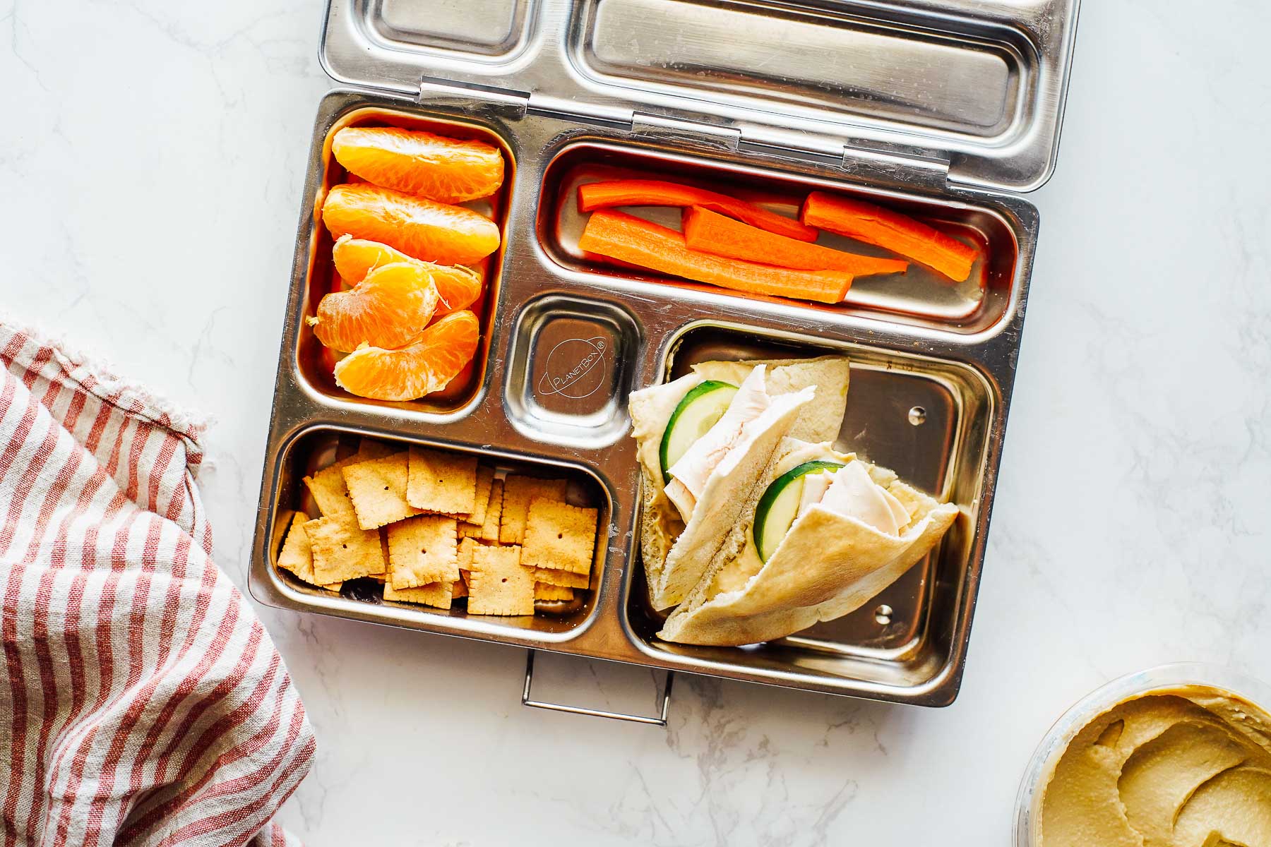 Turkey slices with hummus and cucumber slices in a pita bread, crackers, orange slices, and carrots in a lunchbox.