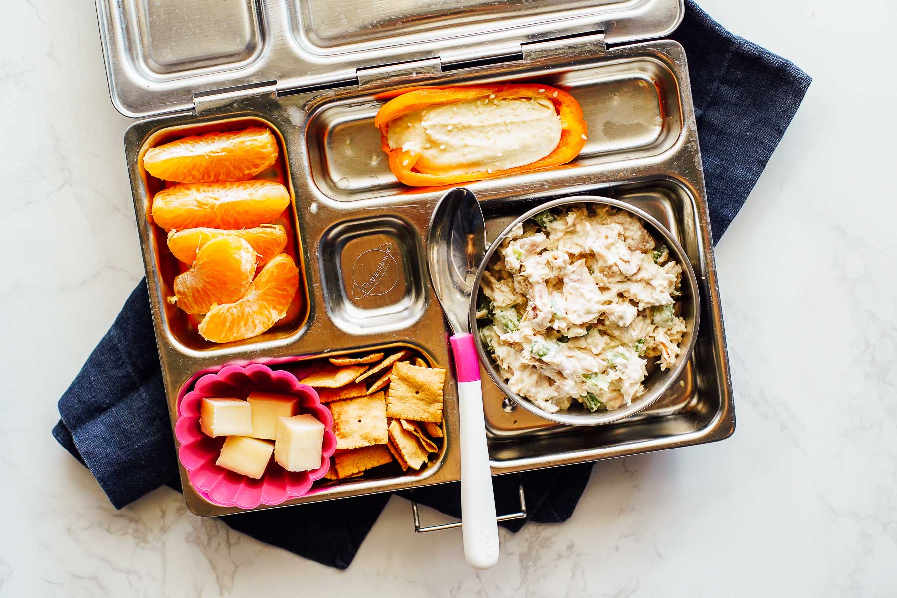 Tuna salad with crackers, cheese cubes, orange slices, and a mini bell pepper with hummus in a bento box.