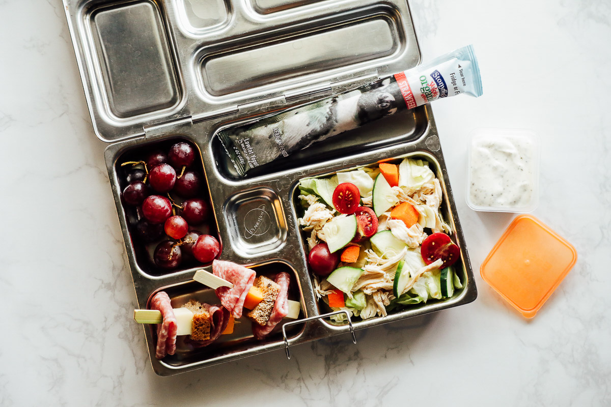 Salad with shredded chicken, salami and bread on skewers, grapes, and a yogurt stick in the lunchbox.