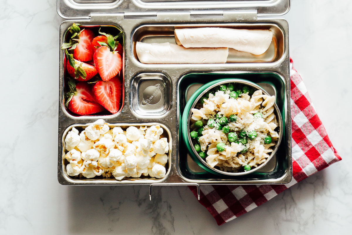 Pasta with peas and parmesan cheese in the lunchbox with rolled up turkey and strawberries and popcorn.