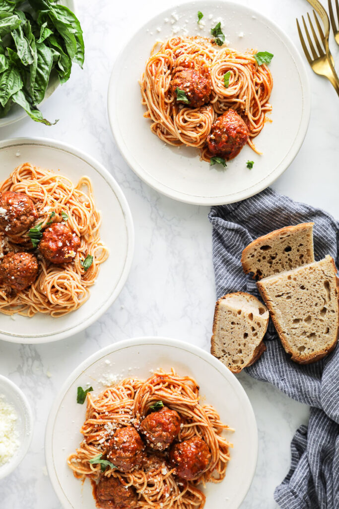 Spaghetti and meatballs plated on 3 white plates with basil and bread on the side.