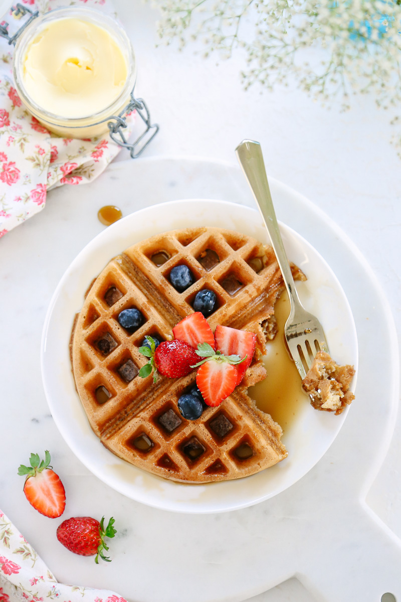 A homemade waffles cut with a fork on a plate with berries and maple syrup on top.