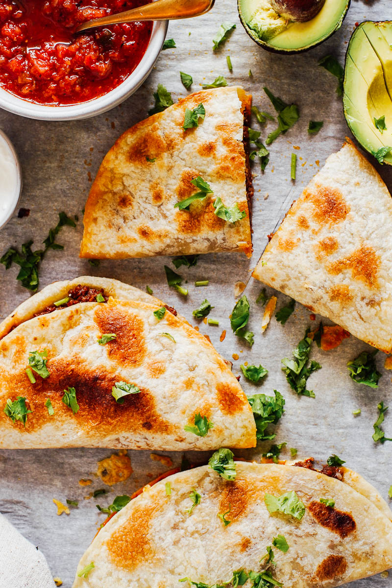 Cooked quesadillas on a sheet pan with salsa and avocados on the side.