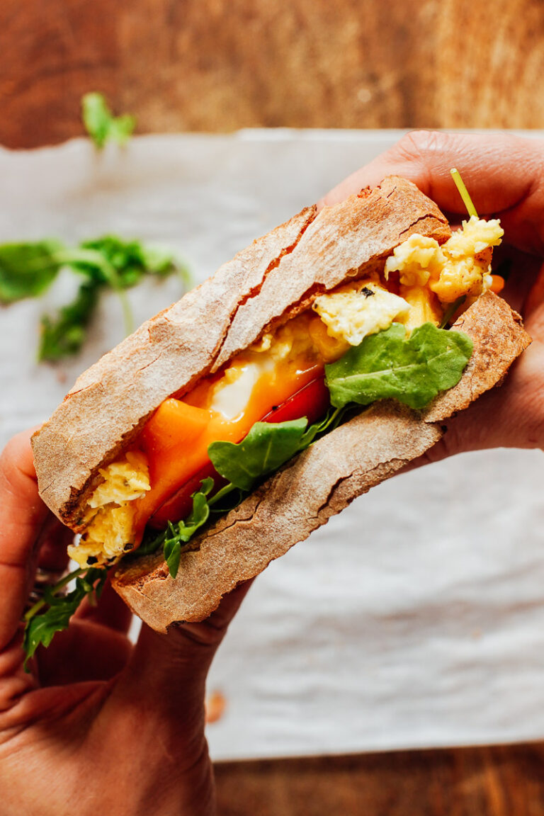 Hands holding an assembled breakfast sandwich: scrambled eggs, cheese, arugula.