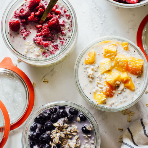 Overnight oats in three jars with frozen fruit on top: mango, blueberries, and raspberries.