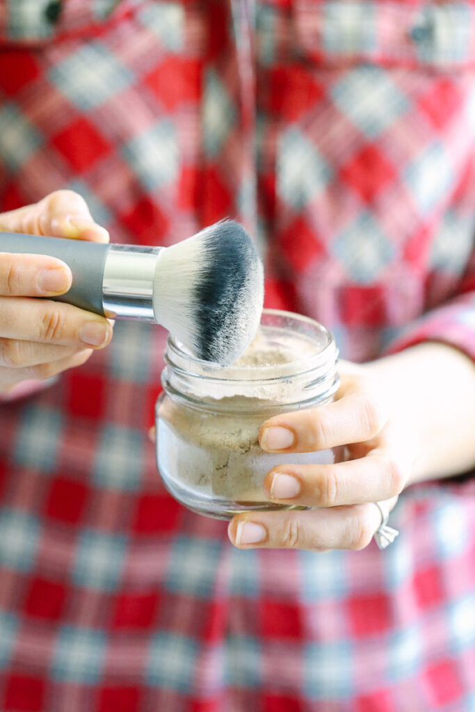 Using a brush to scoop dry shampoo from a glass jar. 